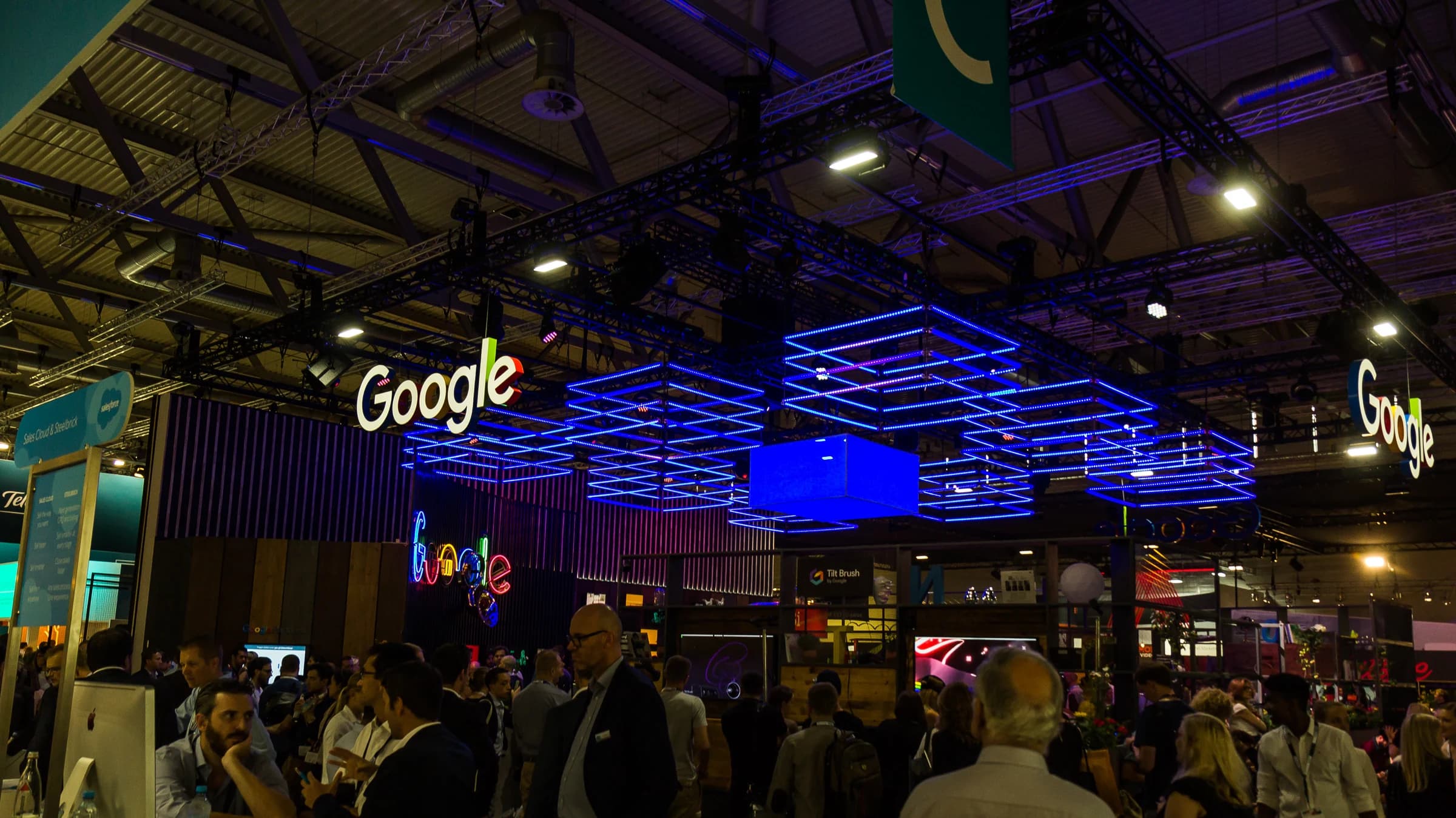 The Google stand at the dmexco digital marketing fair in Cologne, with illuminated Google logos and blue light cubes hanging above a busy crowd.