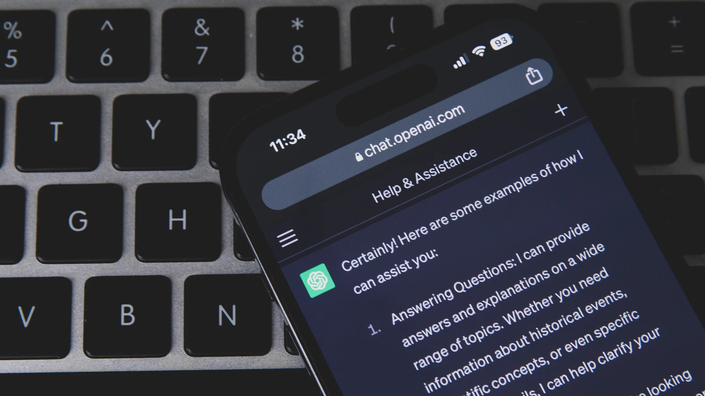 Een smartphone op een laptoptoetsenbord met ChatGPT open in de browser, met een antwoord dat begint met 'Certainly! Here are some examples of how I can assist you'.