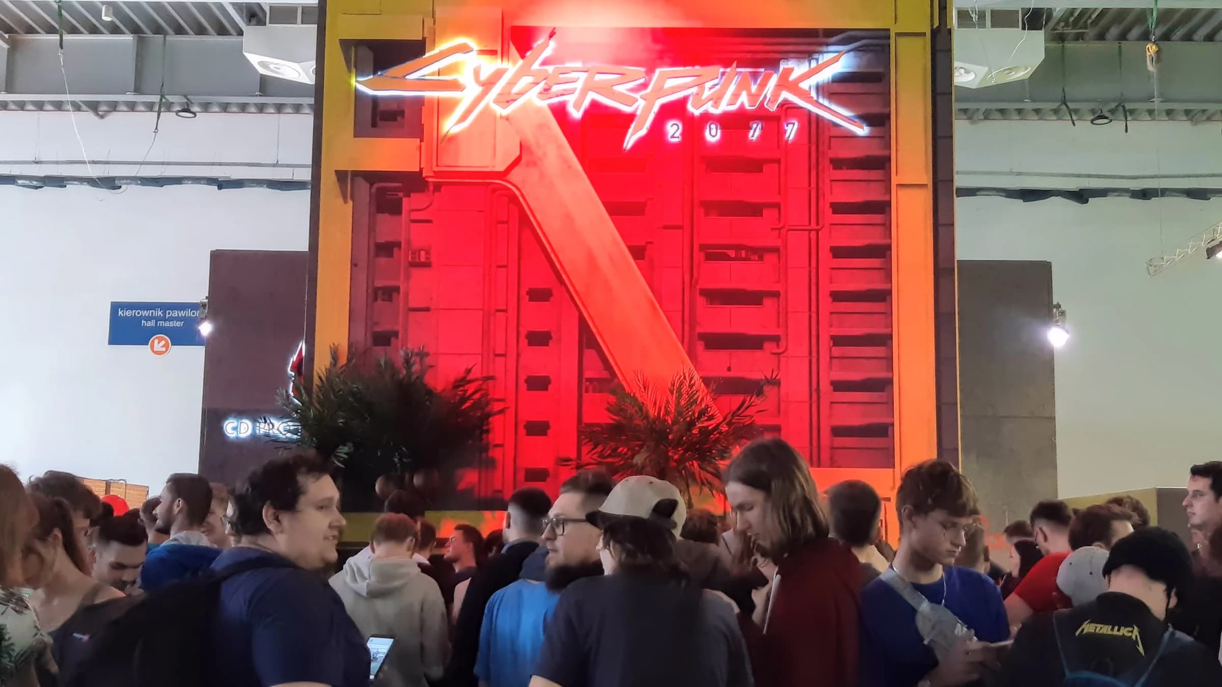 The Cyberpunk 2077 stand at a games fair: the game's logo in red neon above a steel gate, with a crowd of visitors queuing in front of it.