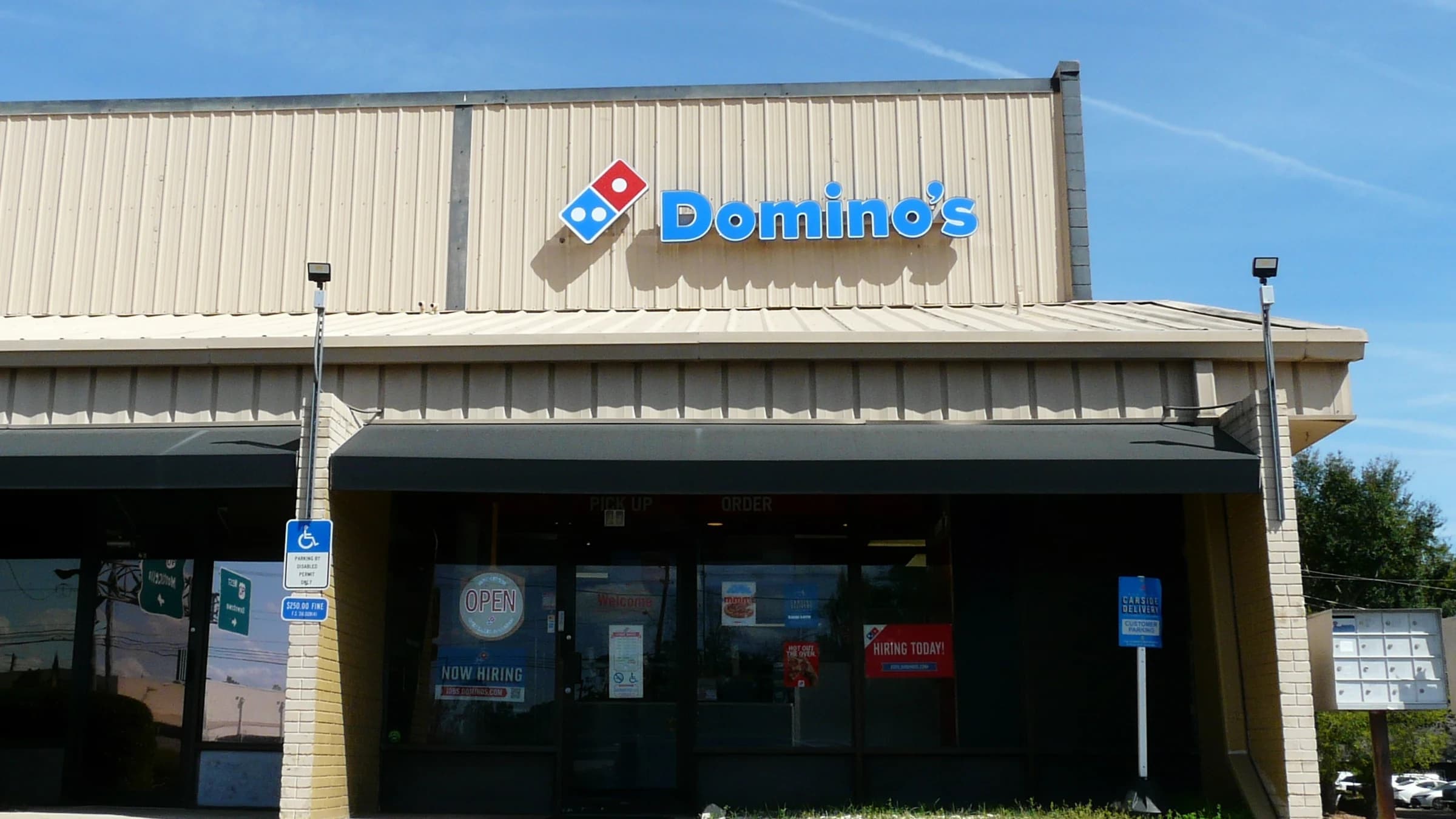 The front of a Domino's store under a blue sky, the logo on the facade above a dark awning and an Open sign in the window.