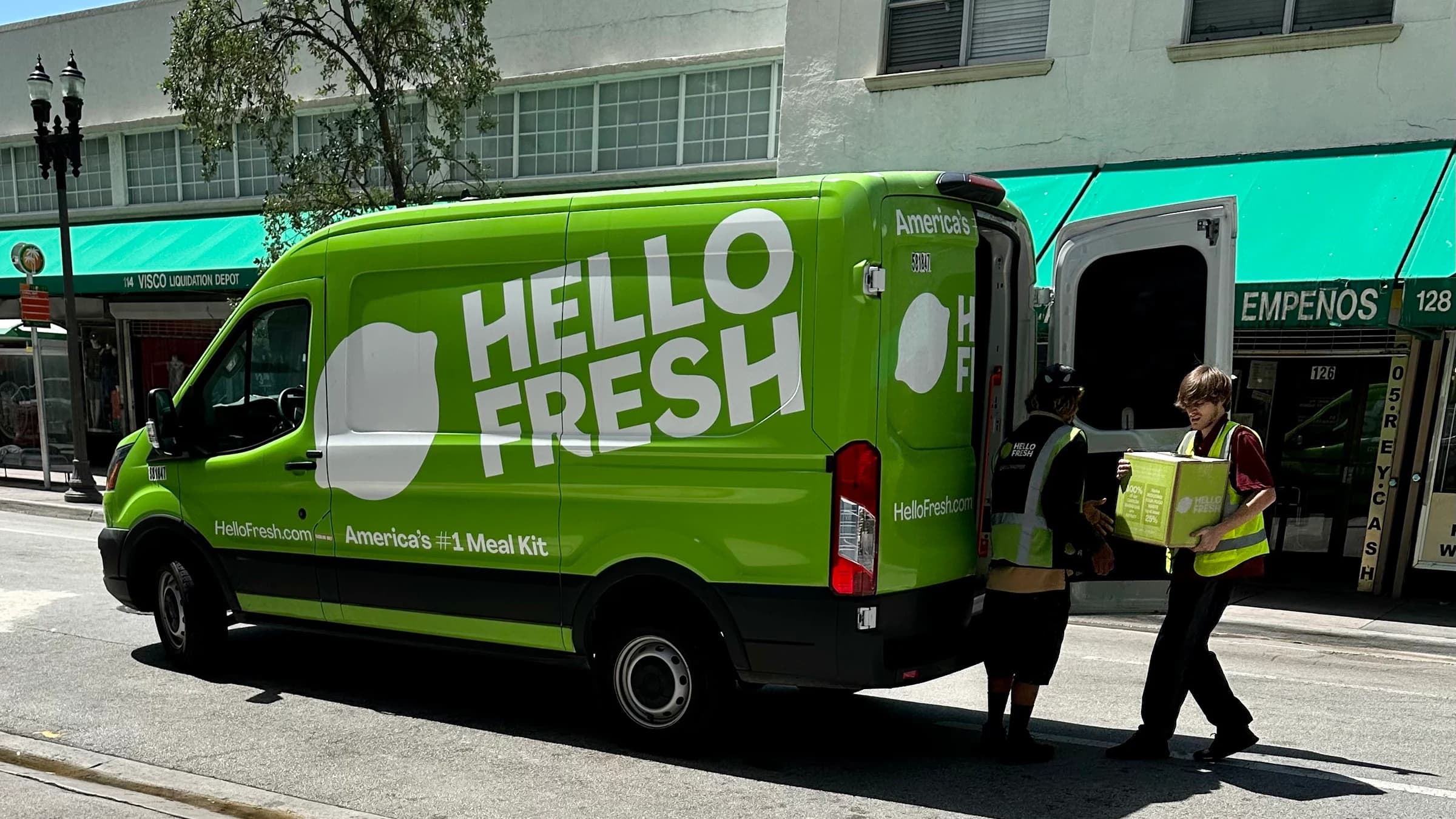 A green HelloFresh delivery van parked on a city street, its back doors open while two couriers unload a meal-kit box.