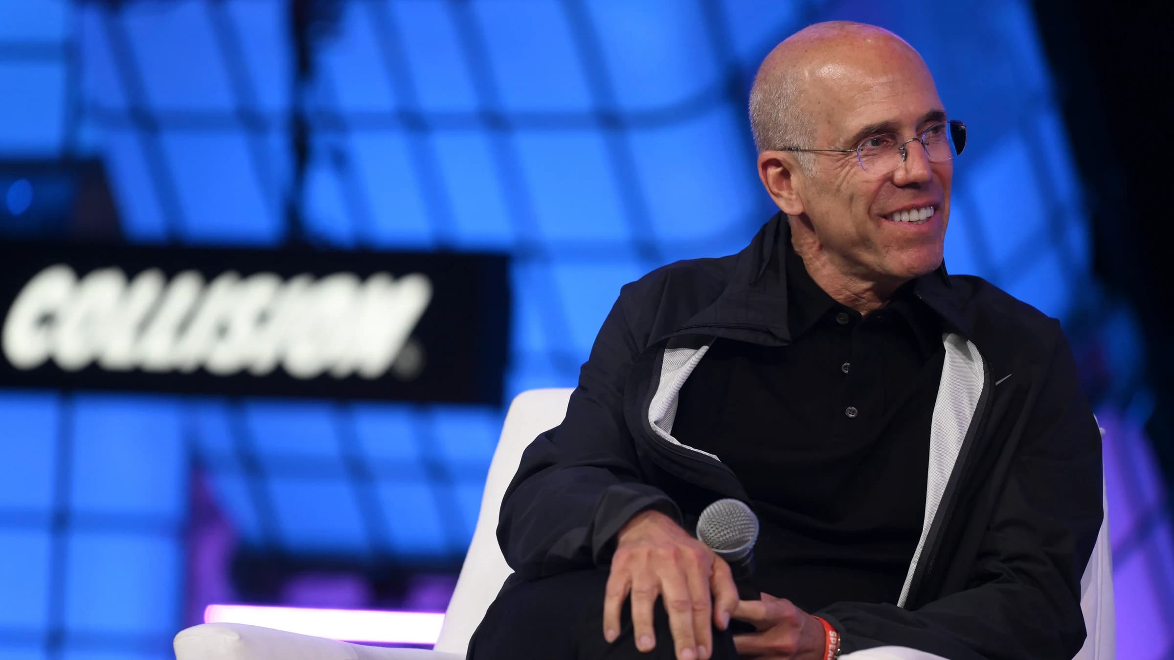 Jeffrey Katzenberg seated on stage in a white chair, holding a microphone, in front of a blue screen at the Collision conference.