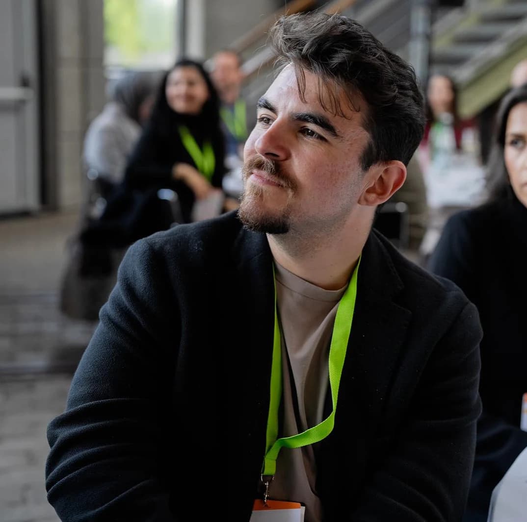 Necati Atakan Alevli in a dark coat and a green lanyard, listening at Diversity Dinner 2025.
