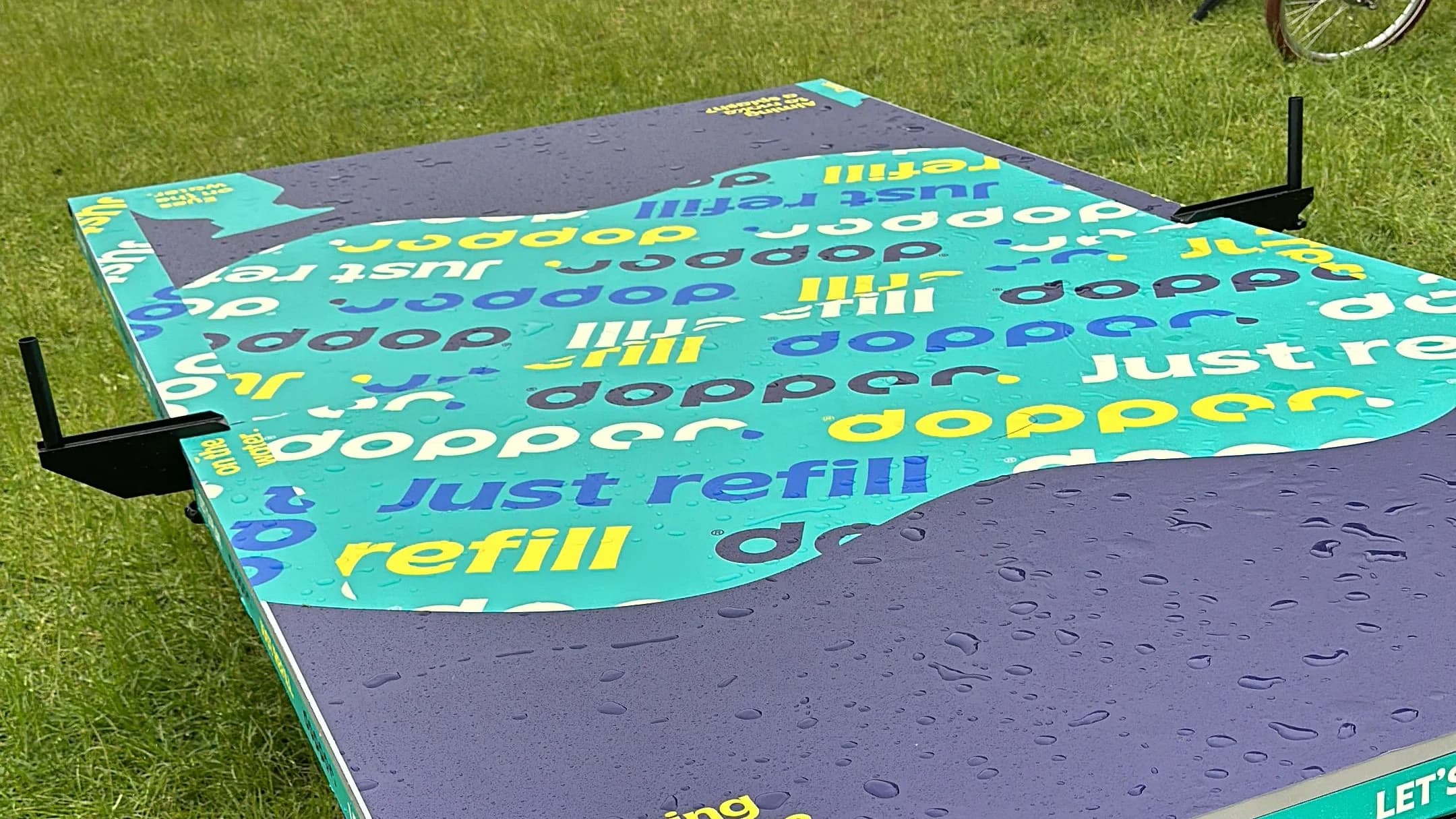 The Dopper Water Pong table set up on festival grass, its top printed in teal, purple and yellow with 'just refill' and 'Aiming to make a splash?', beaded with rain.