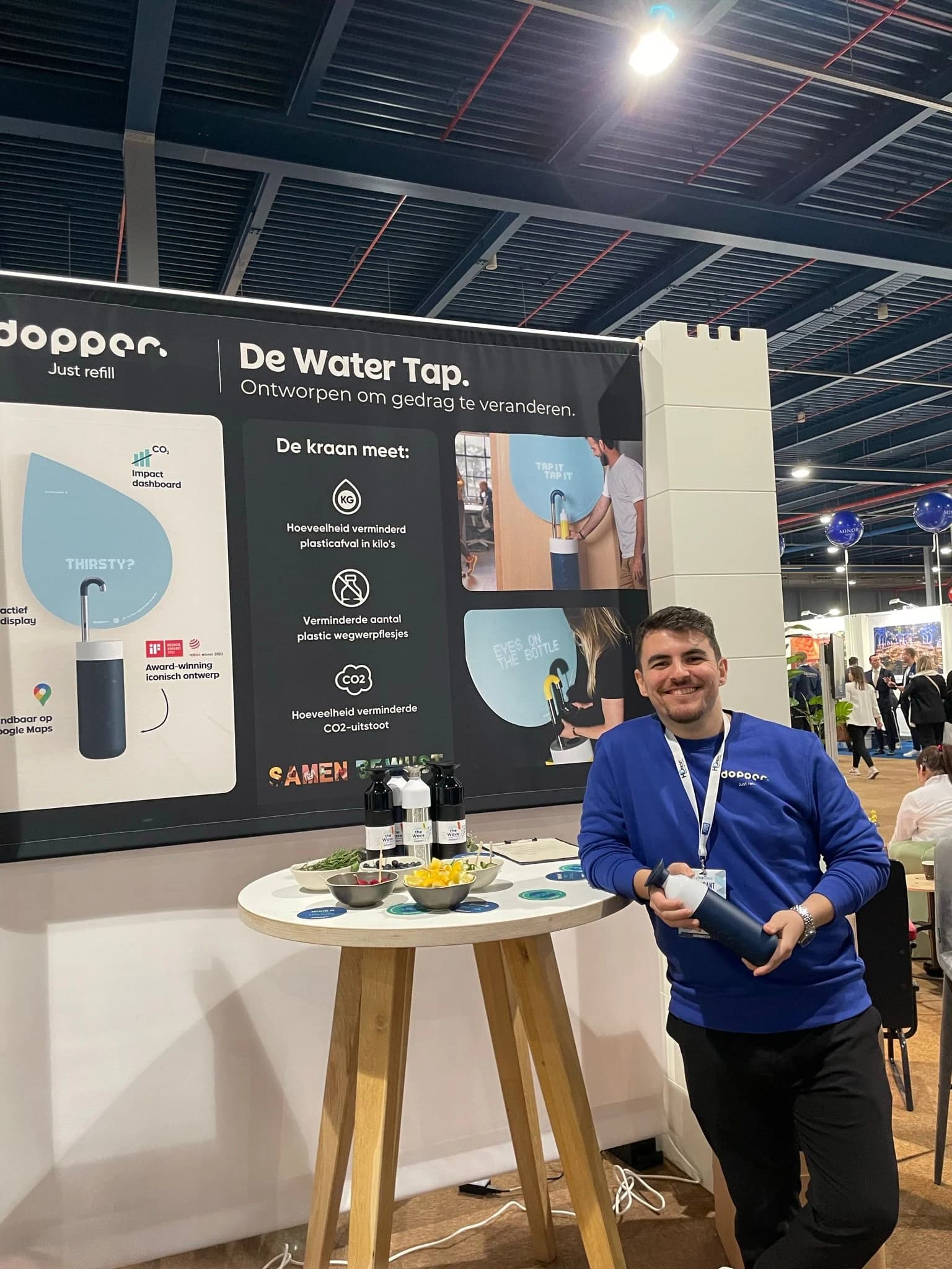 Necati Atakan Alevli in a Dopper sweatshirt beside the Water Tap stand, holding a Dopper bottle.