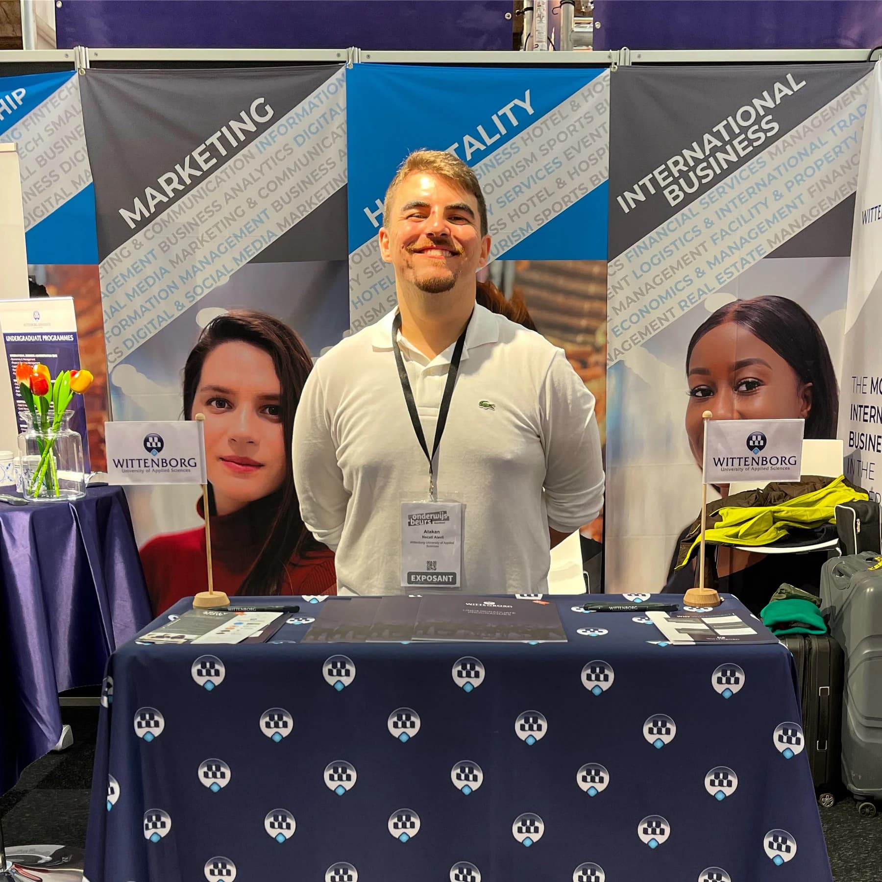 Necati Atakan Alevli behind the Wittenborg stand at an education fair, wearing an exhibitor badge, between two small Wittenborg flags and a table of brochures, with programme banners for Marketing, Hospitality and International Business behind him.