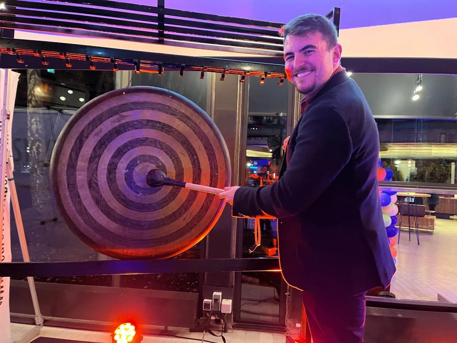 Necati Atakan Alevli in a dark jacket, turning to the camera as he strikes a large wooden gong with a mallet, under orange and purple stage lighting, with orange, white and blue balloons behind him.