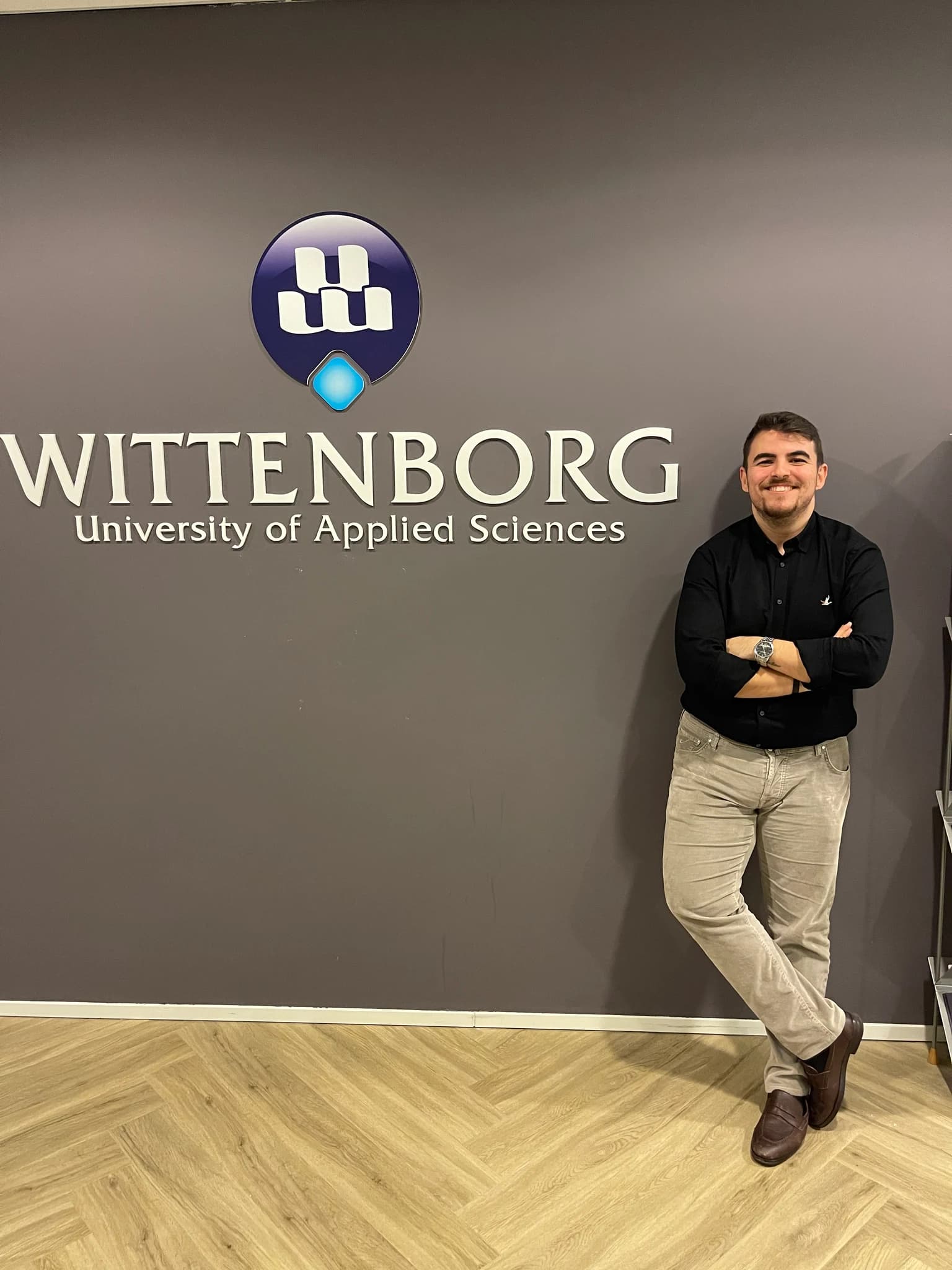 Necati Atakan Alevli in a black shirt, arms crossed, leaning against a grey wall that carries the Wittenborg University of Applied Sciences wordmark and its blue shield mark.